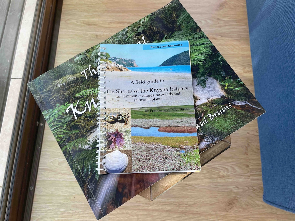 An open field guide is placed atop two larger books, displaying the cover image of the Knysna Estuary. The guide features a spiral binding and includes images of local marine life and plants, providing informative content about the region's ecology.