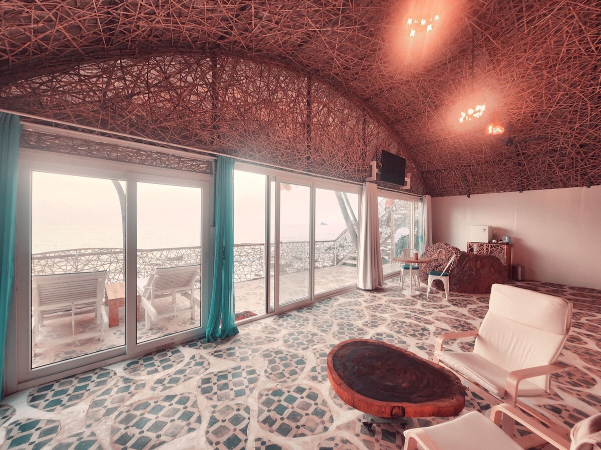 An airy, open space features large glass doors that provide stunning views of the waterfront. The unique, woven ceiling adds an organic texture, while a round wooden table and white lounge chairs are positioned for relaxation. Natural light fills the area, enhancing the tranquil ambiance.