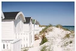 A row of white beach huts is set against a clear blue sky, with the sea visible in the background. The huts feature distinct rooftops, and sandy pathways are framed by dune grass leading to the beach.