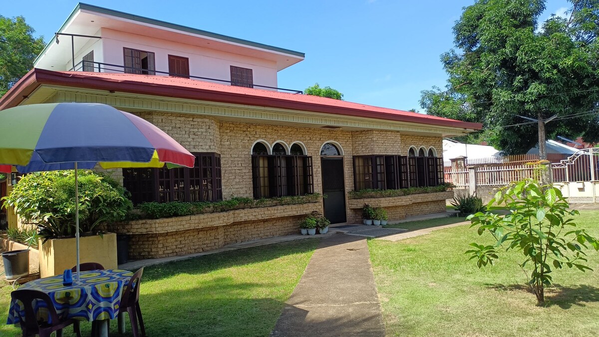 The property features a two-story building with an expansive front lawn. Large arched windows provide ample natural light, while colorful umbrellas offer shade to a seating area. Lush greenery surrounds the outdoor space, creating a serene environment typical of the neighborhood.