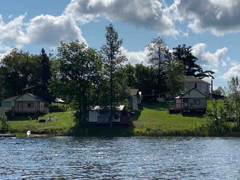 A view of the waterfront accommodations features several cottages set on a grassy slope. Lush trees surround the properties, which are situated against a backdrop of clouds and blue sky, enhancing the natural setting along the water's edge.