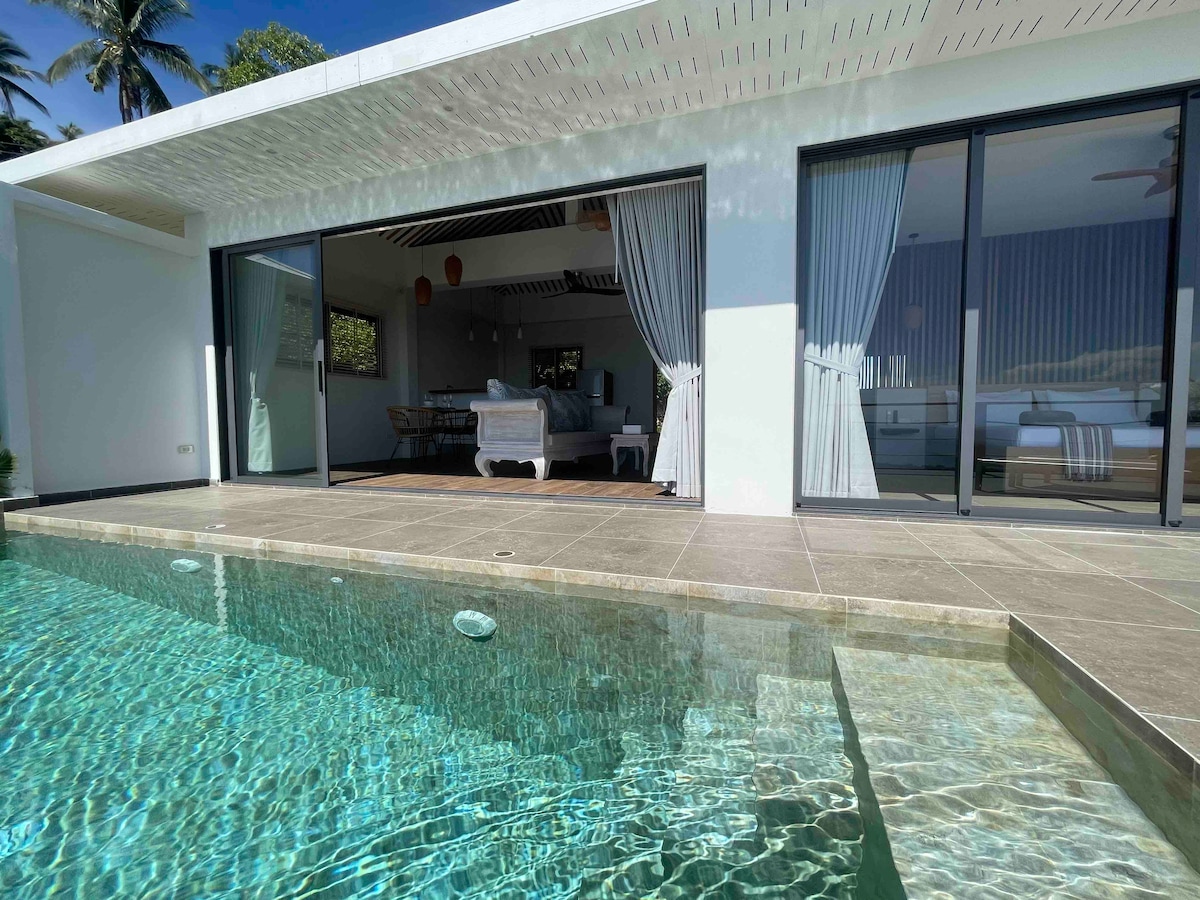A modern villa features expansive sliding glass doors that open to a private infinity pool. The clear water reflects the sunlight, while a spacious outdoor area is visible, seamlessly connecting the interior living space with nature.