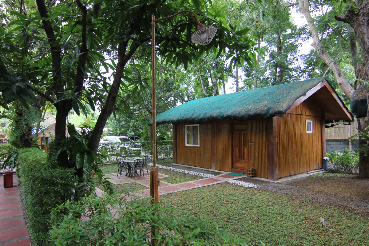 A cozy cottage is framed by lush greenery, featuring a thatched roof and wooden siding. A small yard is visible with a stone path and outdoor dining area, surrounded by manicured grass and shaded by trees. The inviting entrance includes a single window with white framing.