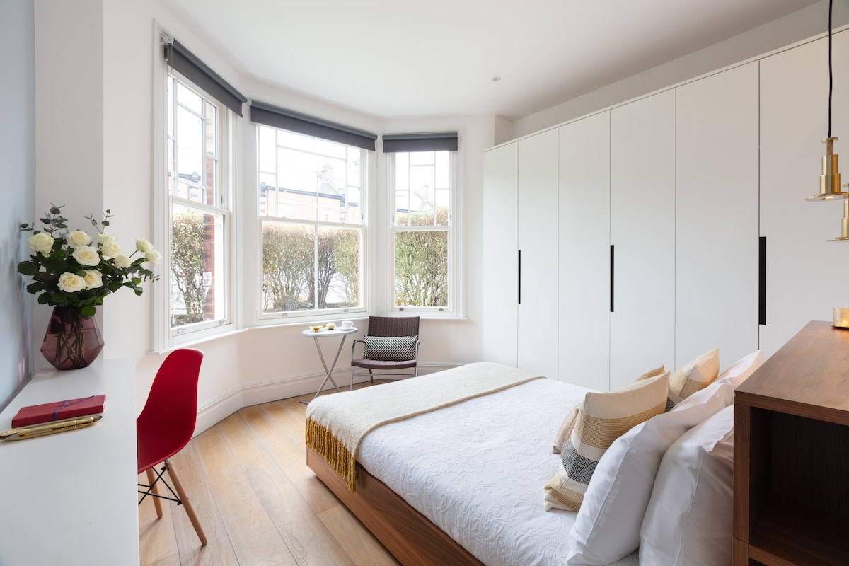 The bedroom features a spacious bed with neutral bedding and decorative throw. Large bay windows provide natural light, partially covered by blinds. A compact seating area includes a chair and small table, while built-in cabinetry offers storage space. A vase of flowers adds a touch of greenery.