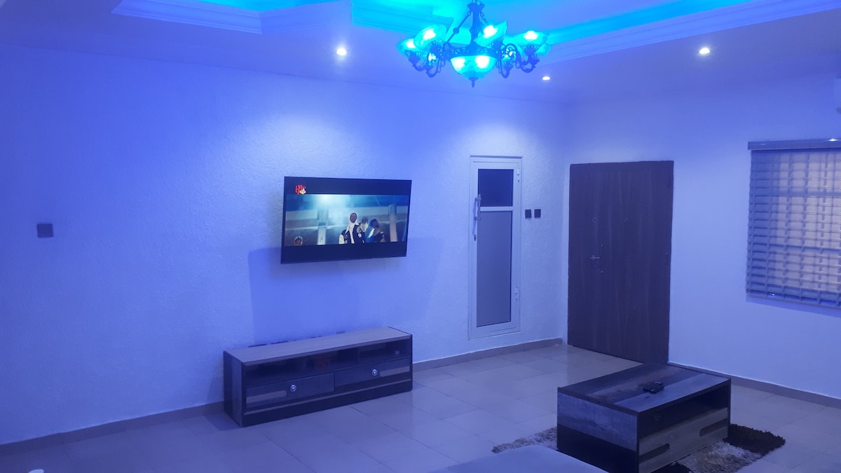 The living room features a modern design with soft blue lighting enhancing the space. A wall-mounted flat-screen TV displays programming, while a low coffee table sits on a light-colored area rug. A door, window, and decorative wall elements complete the inviting atmosphere.