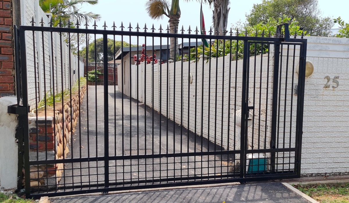 A secure entrance features a black metal gate with vertical bars, flanked by a stone wall. A paved driveway is visible, leading to the residence. Lush greenery and palm trees are seen in the background, enhancing the welcoming ambiance of the property.