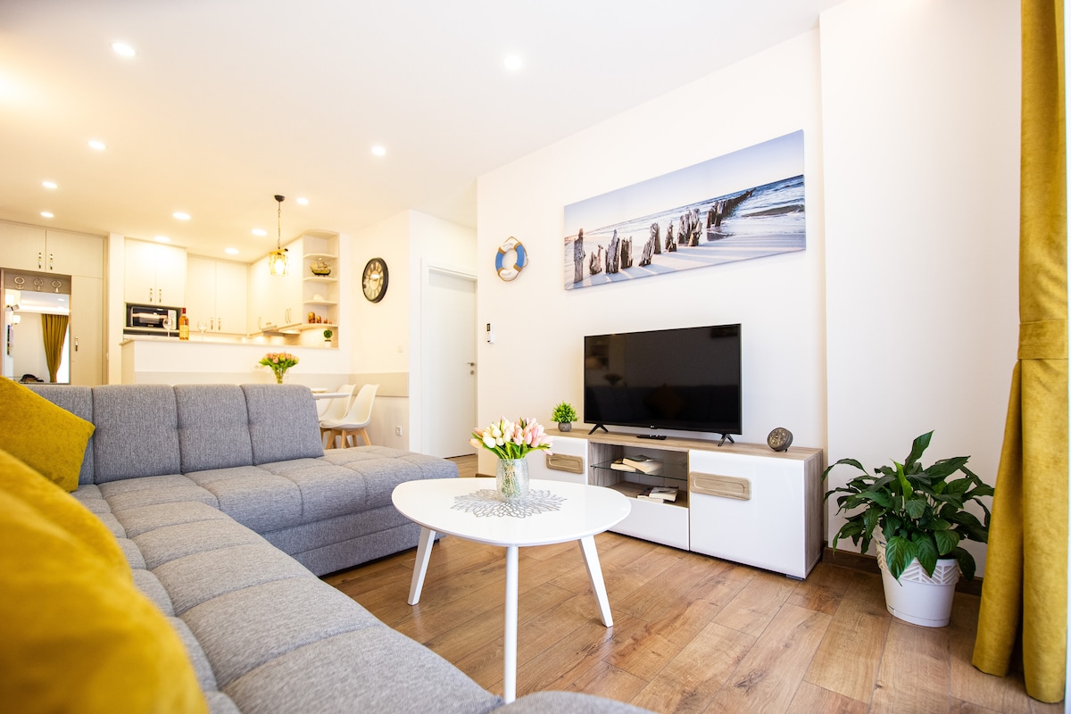 A modern living area features a large gray sectional sofa with yellow accent pillows. A white coffee table sits in front of a media console below a mounted television. Light wood flooring complements the space, illuminated by recessed ceiling lights and natural light from the nearby kitchen.