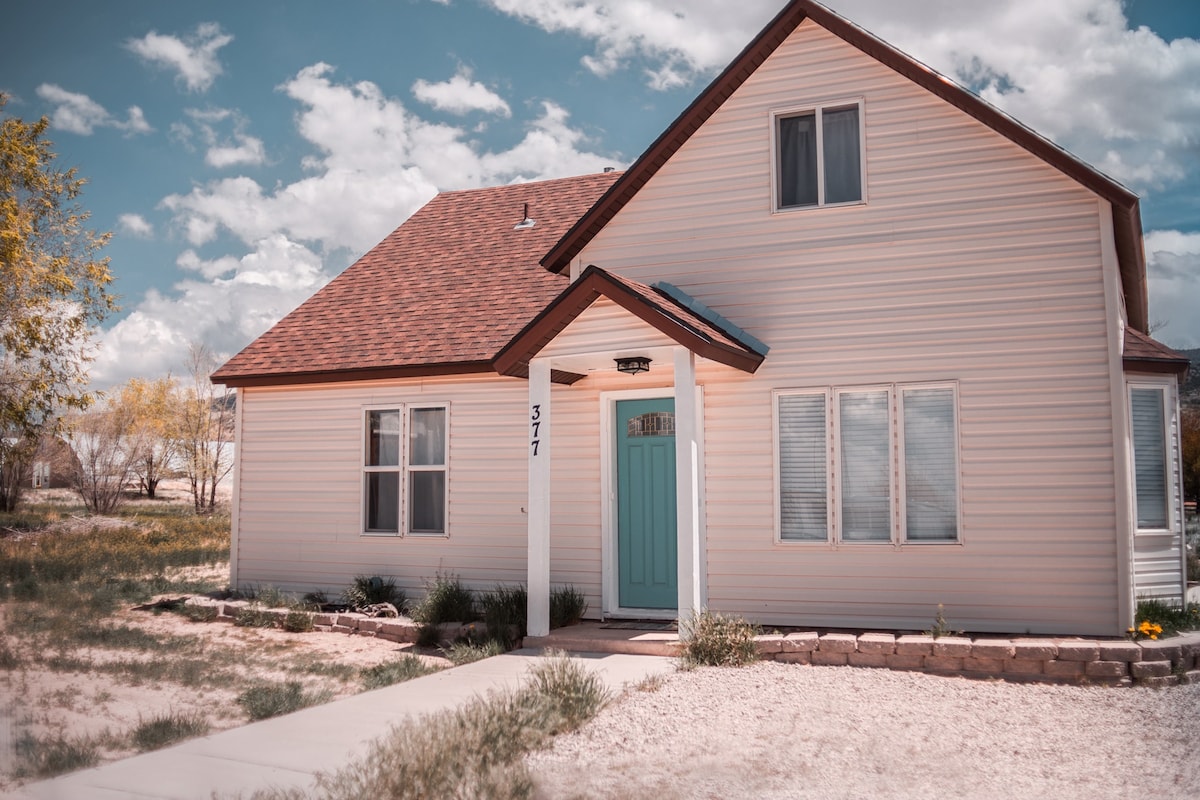 The exterior of a two-story home is depicted, featuring a light-colored facade with a sloped roof. A welcoming blue door is central, flanked by large windows. The pathway is lined with simple landscaping, set against an expansive sky with scattered clouds.