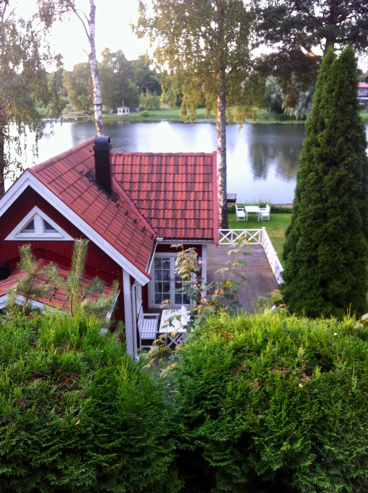 A charming red villa with a sloping tiled roof is nestled among greenery. The terrace extends towards a serene lake, framed by tall trees. Outdoor seating is visible on the terrace, providing a tranquil view of the water.