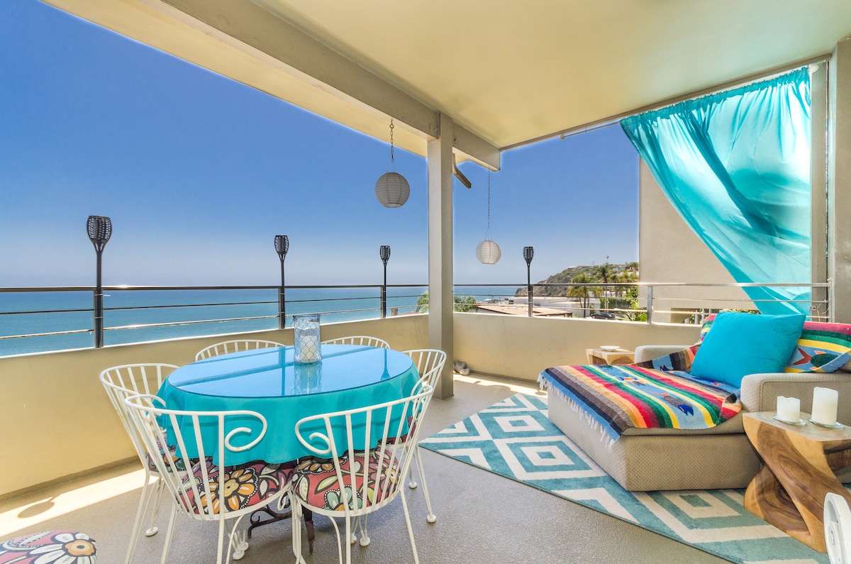 A large private balcony features a round dining table with colorful chairs, overlooking unobstructed ocean views. A cozy lounge area and decorative pillows are positioned beside a patterned rug. Soft curtains sway gently in the breeze, enhancing the tranquil atmosphere of this oceanfront space.