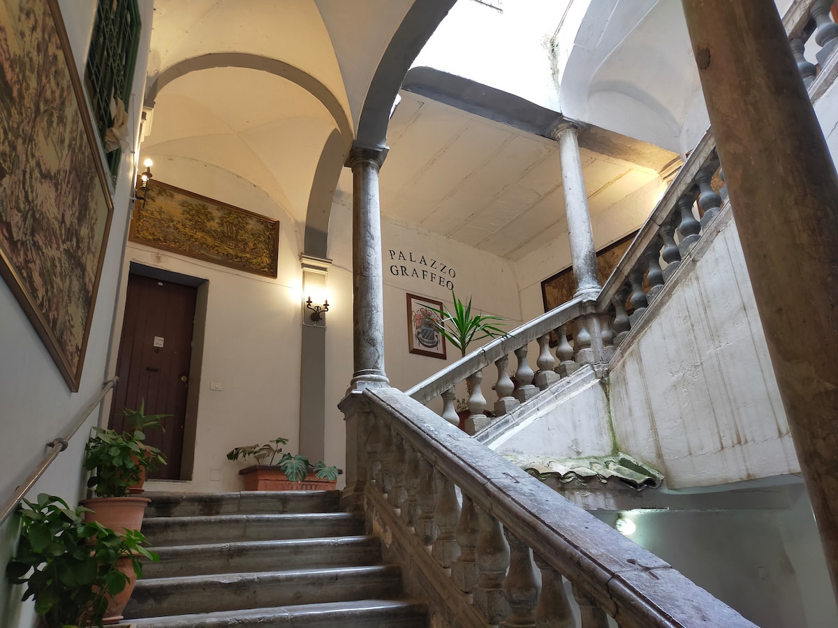 An elegant staircase with a curved stone railing leads to an upper level. Potted plants are placed along the steps, and decorative wall art adds character to the space. Light filters softly through the openings above, enhancing the ambiance of the centrally located building.