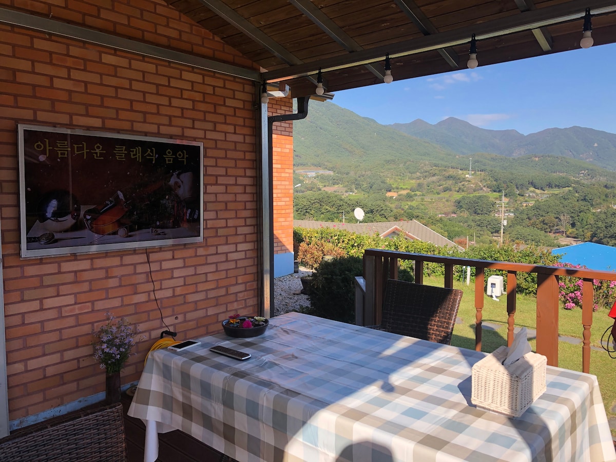 An outdoor dining area features a wooden table set against a backdrop of lush mountains. A framed picture hangs on the wall, and decorative lighting is visible above. The expansive view reveals green hills under a clear sky, creating a peaceful atmosphere.