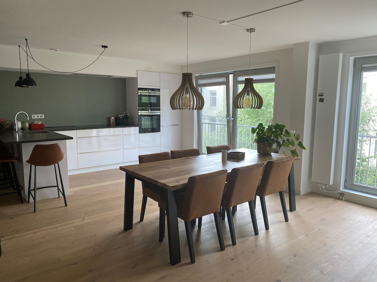An open kitchen and dining area is highlighted by a large wooden table surrounded by brown chairs. Modern light fixtures hang above, and expansive windows allow natural light to fill the space. The kitchen is equipped with sleek, white cabinetry and stainless appliances.