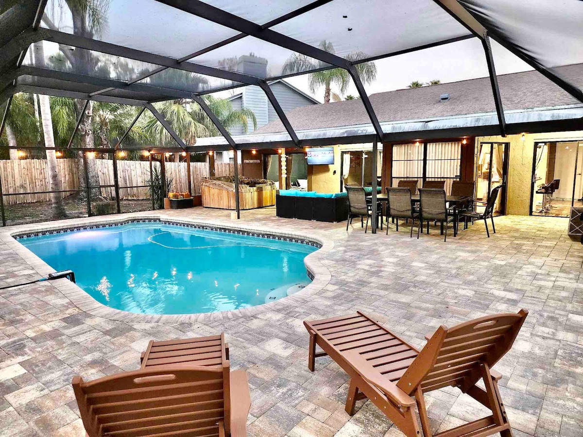 An outdoor space features a screened-in area with a sparkling pool surrounded by paver stones. Lounging chairs are positioned nearby, overlooking the pool. A hot tub and outdoor seating area with tables are also visible, providing options for relaxation and socializing.
