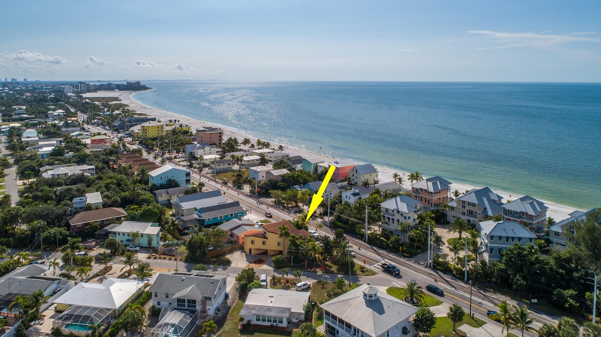 An aerial view showcases the proximity of the condo to Fort Myers Beach, highlighting the coast and nearby buildings. The beach stretches along the horizon, with the residence positioned in the foreground, visibly adjacent to Estero Boulevard.