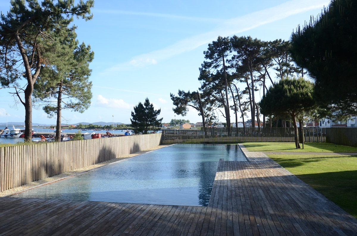 A serene swimming pool is set within a wooden deck, surrounded by well-maintained grass and tall trees. The clear water reflects the sky, with distant views of boats in the harbor beyond the wooden fence.
