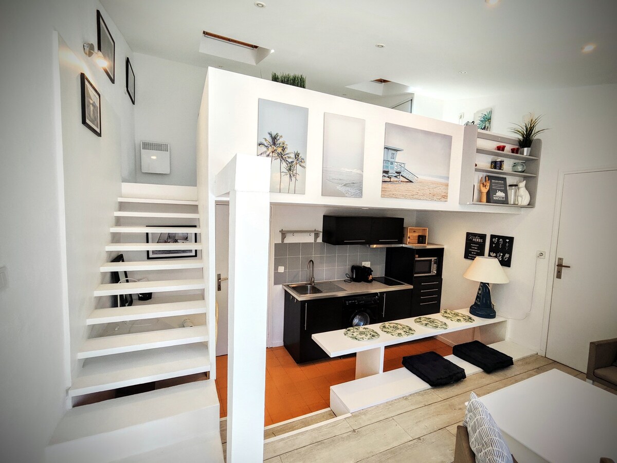 An open living area features a modern kitchen with a sleek countertop and ample shelving. Stairs lead to a mezzanine bedroom area. Decor includes beach-themed artwork displayed on white walls, enhancing the light and airy aesthetic. Comfortable seating and dining options are arranged below.