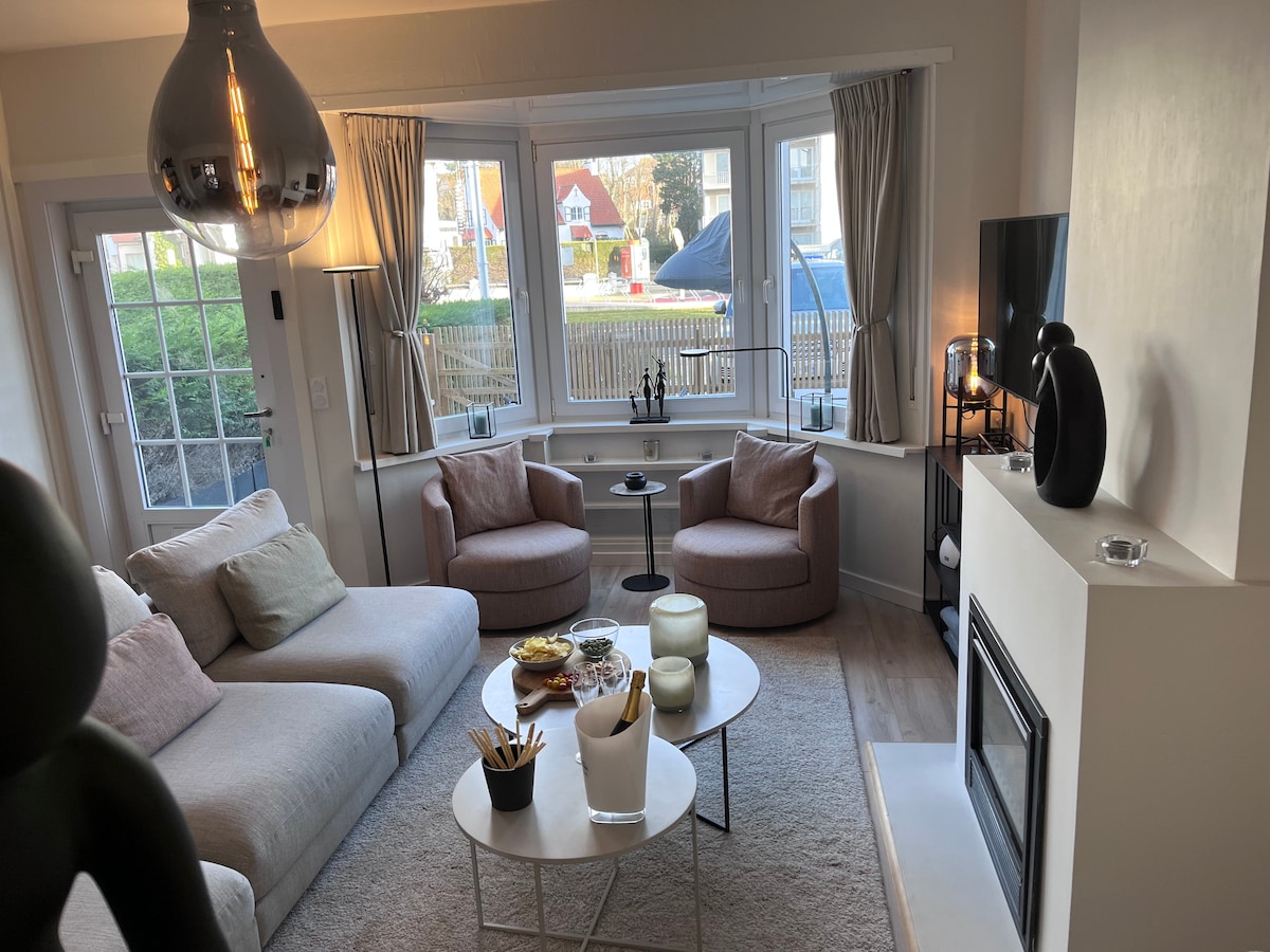 A well-lit living area features a comfortable sofa and two lounge chairs, inviting relaxation. A modern coffee table holds decorative items, while a fireplace adds a cozy touch. Large windows provide natural light and views of the outside, enhancing the open and airy feel.