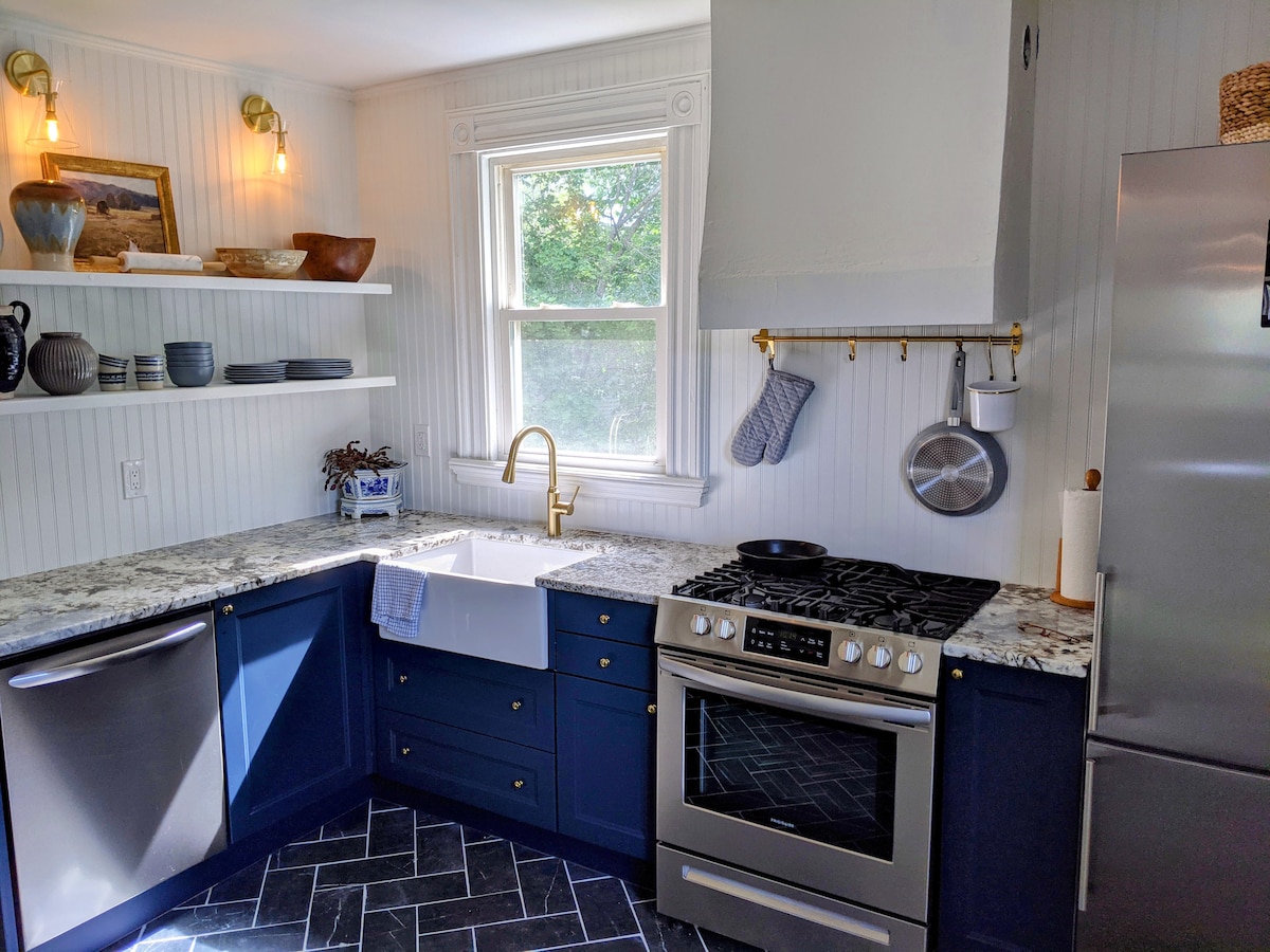 A well-equipped kitchen features blue cabinetry and white walls, complemented by granite countertops. A farmhouse sink is positioned beneath a window, allowing natural light to enter. Shelves display decorative bowls and cookware, while a gas stove occupies one corner, enhancing functionality.