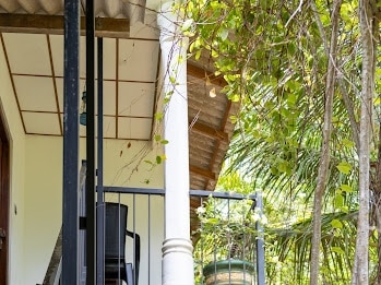 A balcony is visible, featuring a relaxed seating arrangement with a chair. Lush greenery surrounds the area, providing a natural ambiance. The structure showcases a traditional roof design and is complemented by decorative pillars.