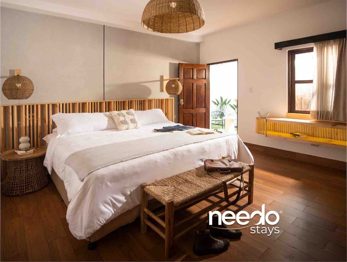 The sleeping area features a spacious bed with a light-colored duvet and decorative pillows. Natural light enters through a window adorned with sheer curtains. A wooden headboard complements the earthy tones of the room, which includes a woven basket and a wooden chair.