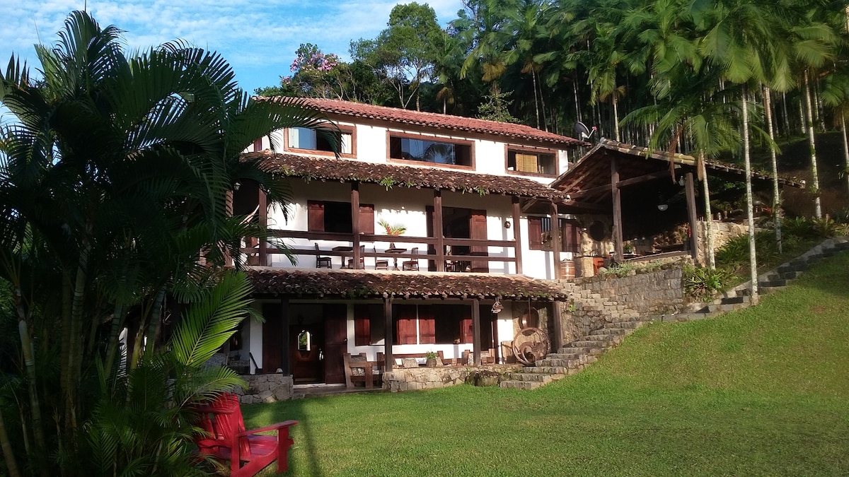 A charming chalet is featured, nestled amid lush green surroundings. The two-story structure showcases wooden balconies and a terraced garden. Various seating areas are arranged on the porch, surrounded by palm trees, creating an inviting outdoor space.
