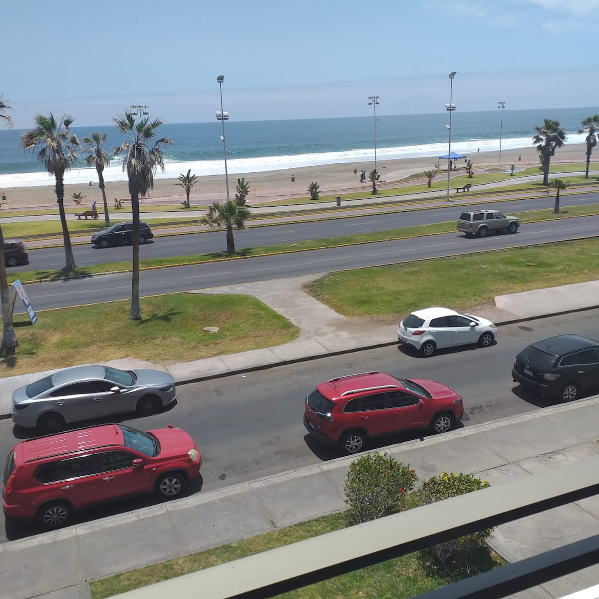 A view from the balcony shows a wide, sandy beach with gentle waves lapping at the shore. Palm trees line the beachfront, while a pathway is visible for strolling. Several parked cars are situated along the street in front, indicating a lively atmosphere.