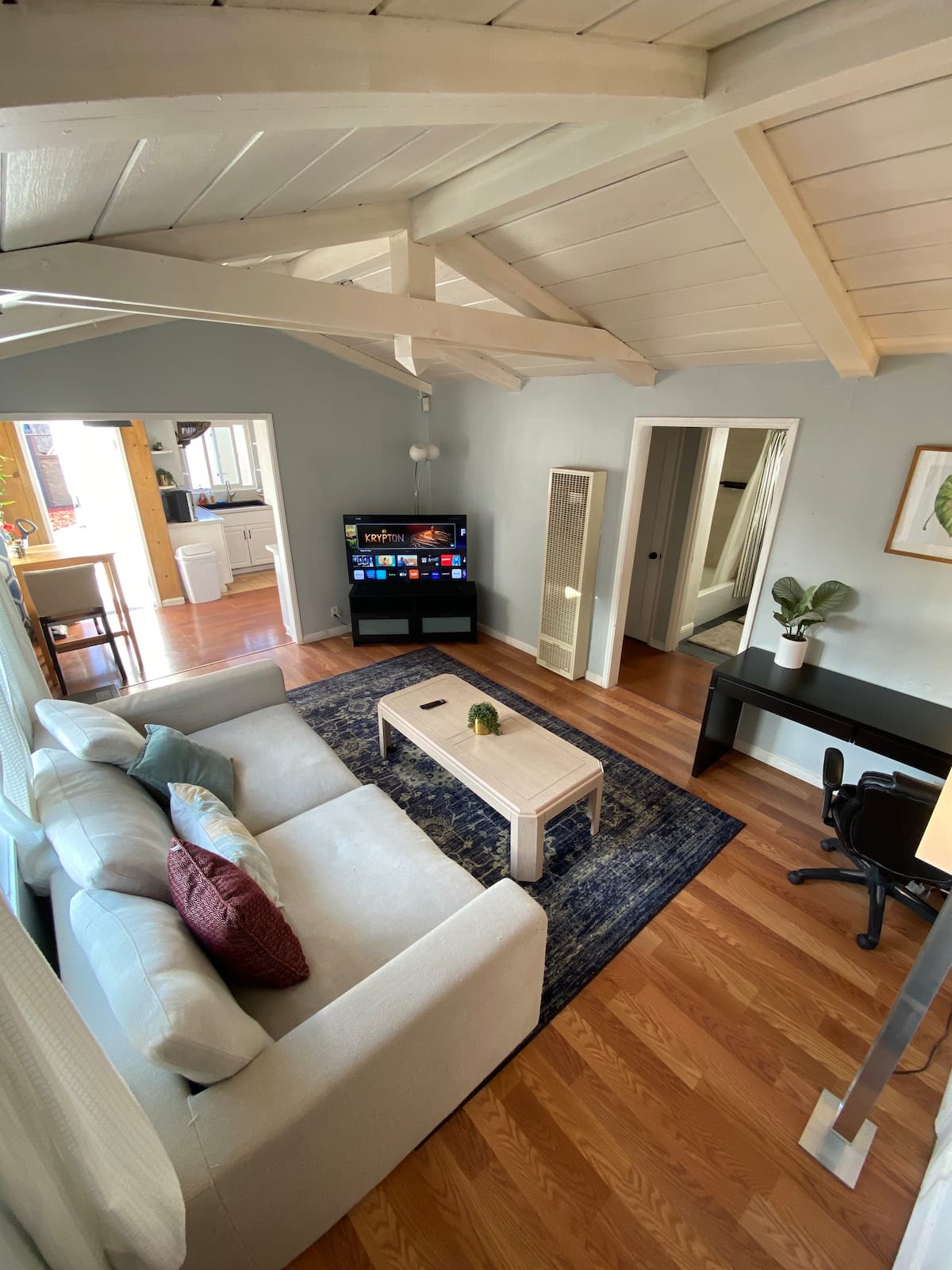 A cozy living area is highlighted by a light-colored sectional sofa adorned with decorative pillows. A wooden coffee table sits in front of the sofa, while a flat-screen TV is positioned nearby. Wood flooring and vaulted ceilings create an open and airy feel, with natural light enhancing the space.