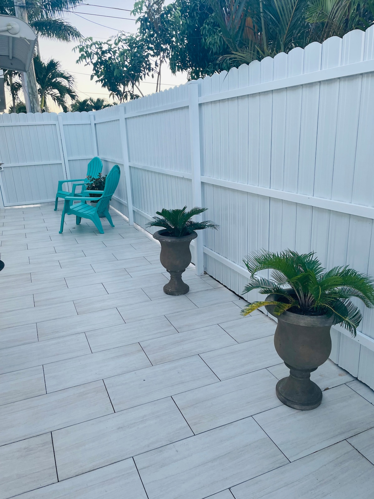 A private outdoor space is highlighted by white fencing and tiled flooring. Two decorative planters with greenery are positioned beside the walkway, and a turquoise chair adds a pop of color, inviting moments of relaxation.