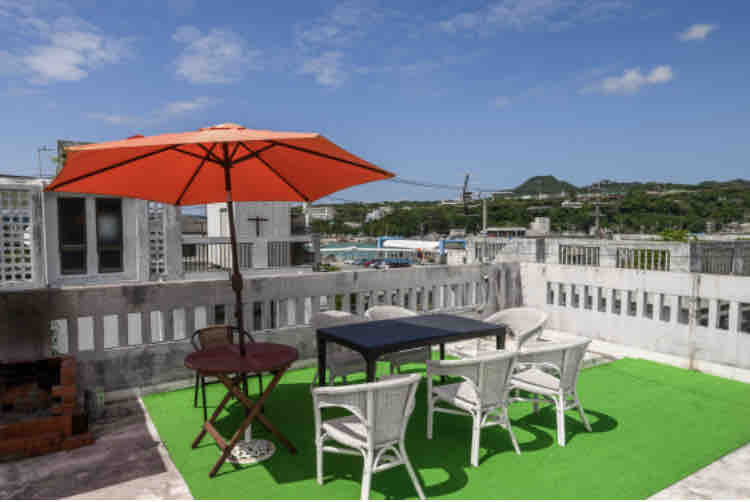 A rooftop space is presented with a vibrant green surface, featuring a round table and four white chairs. An orange umbrella provides shade, while a brick barbecue grill is visible in the background. Scenic views can be seen in the distance, enhancing the outdoor experience.