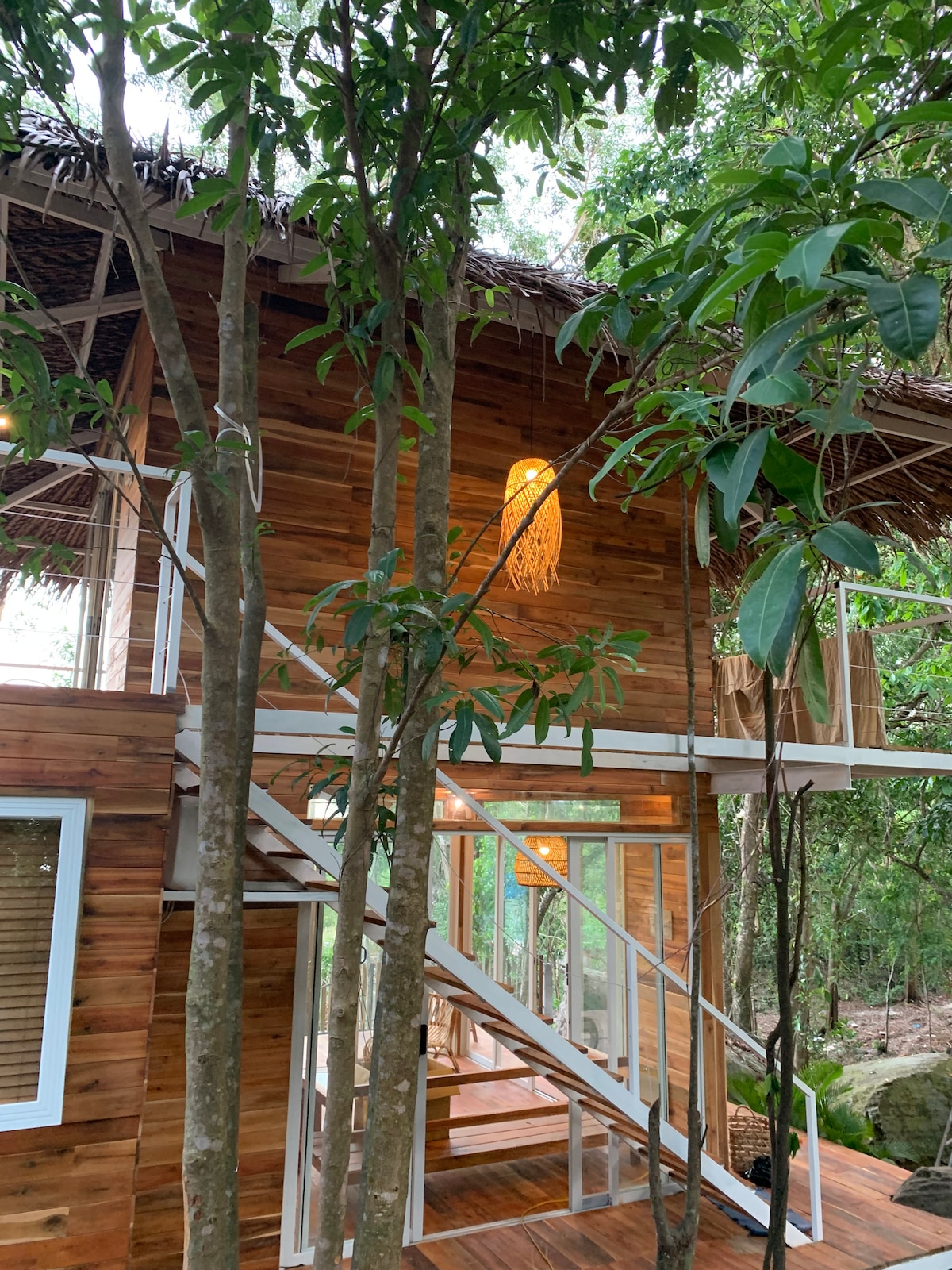A timber house stands at the edge of a forest, surrounded by trees. A staircase leads to the upper level. The exterior features wood paneling and a thatched roof. Soft lighting creates a warm ambiance, enhancing the connection with nature.