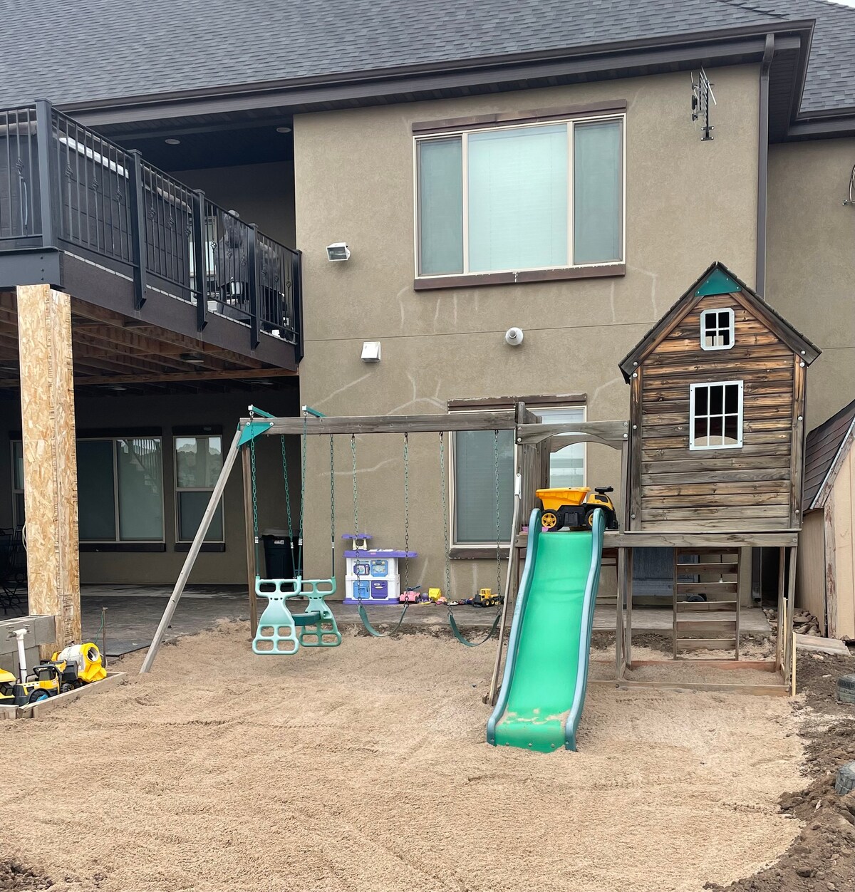 A playset featuring a wooden house structure with a slide and swings stands in a sand-covered yard. Bright green equipment is visible alongside toys, and the backdrop includes the exterior of the nearby building with large windows.
