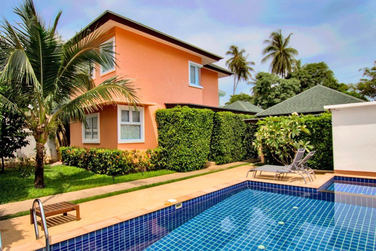 A two-story villa is surrounded by lush greenery, with vibrant orange exterior walls. A shared swimming pool is visible in the foreground, featuring blue tiles and a comfortable lounging area with two sun loungers. Palm trees provide a tropical ambiance.