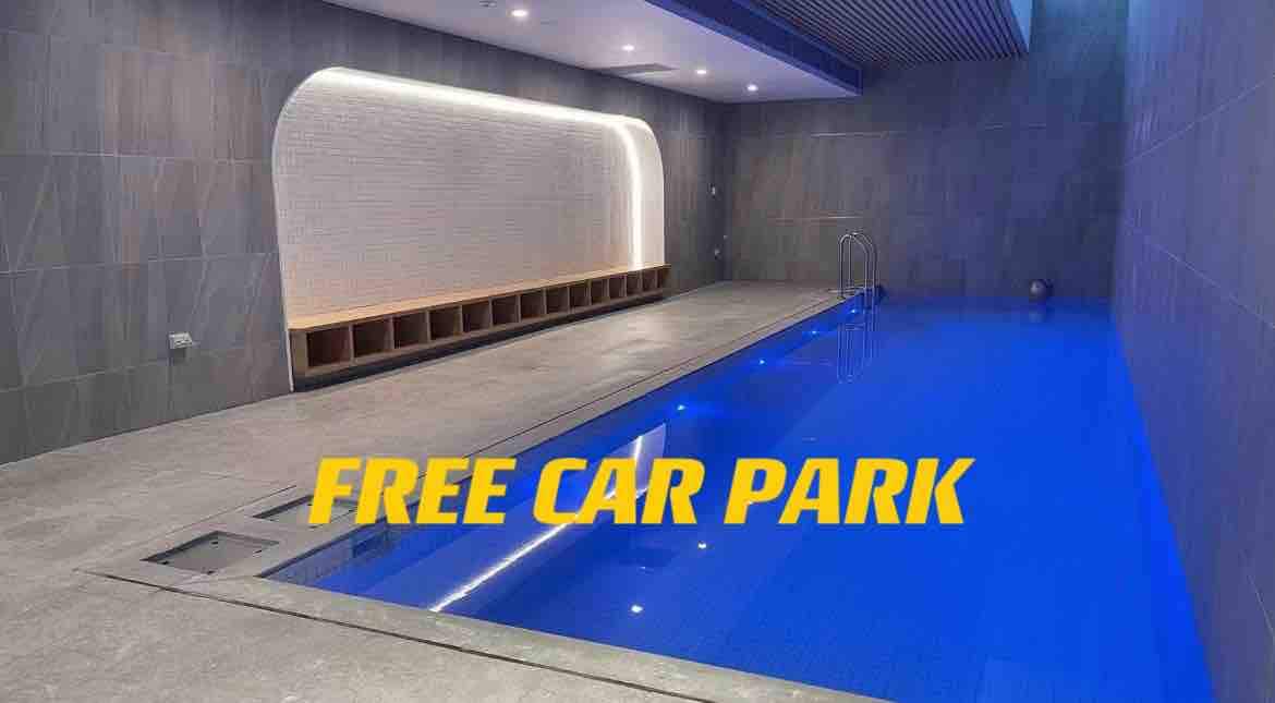 An indoor swimming pool is presented with blue water and sleek tile walls. Natural light is emitted through recessed lighting along the ceiling. A wooden bench is positioned beside the pool, providing seating for relaxation.