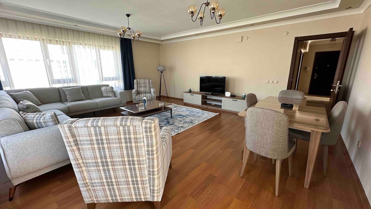 A spacious living area features a large, comfortable sectional sofa and two armchairs with a patterned design. A wooden table is set for dining beside a sleek entertainment unit and a flat-screen TV. Natural light fills the room through large windows with sheer curtains.