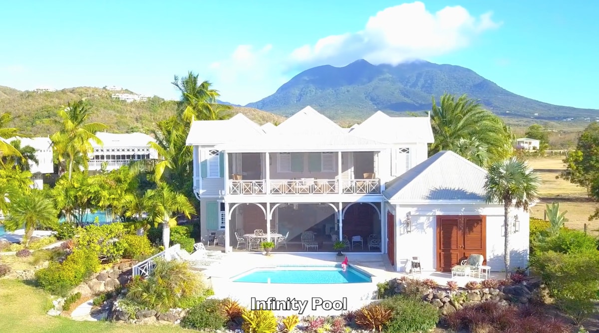 An aerial view showcases a luxury villa with a private infinity pool surrounded by lush landscaping. The backdrop features a prominent mountain, while the villa displays a welcoming design with balconies and outdoor seating areas.
