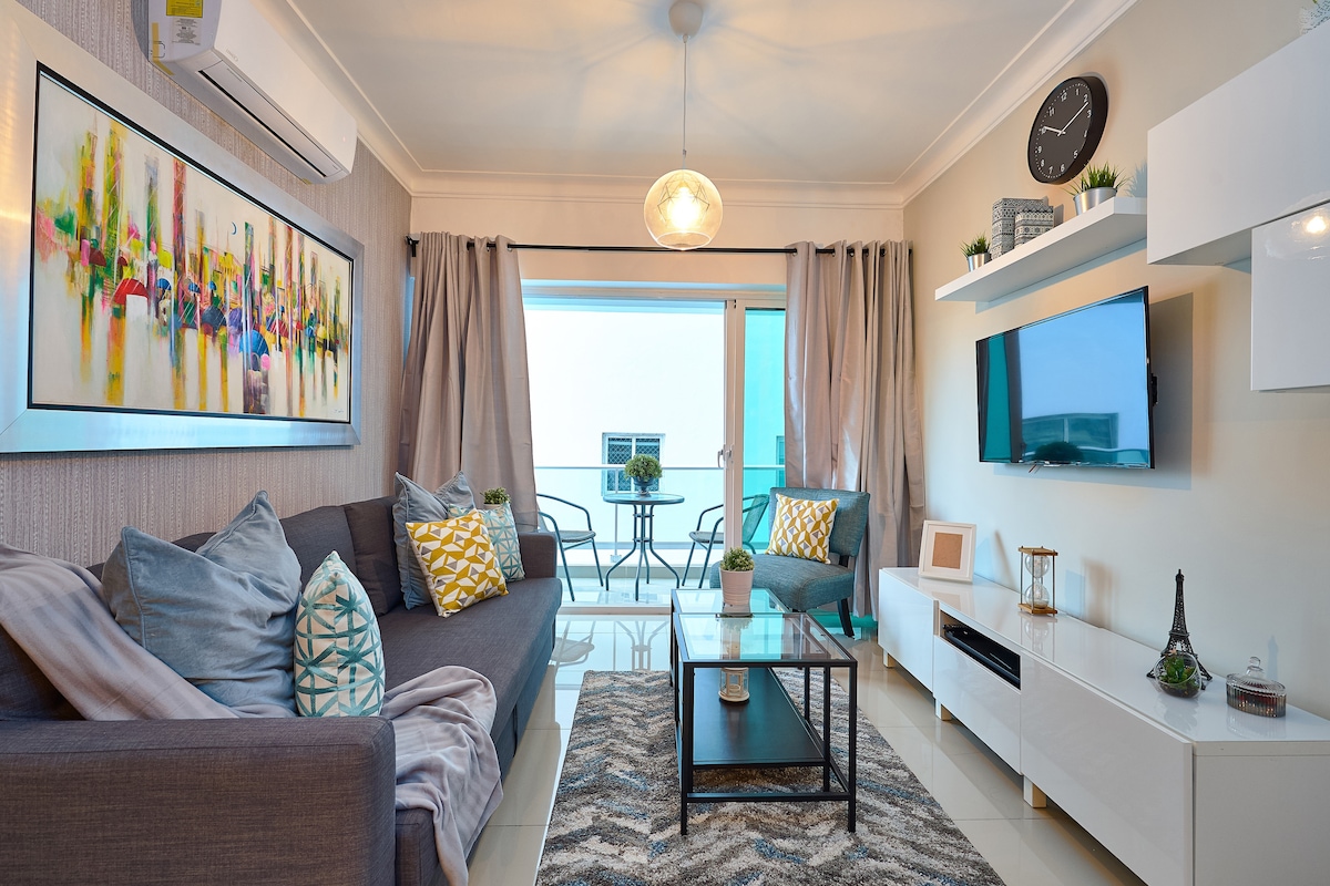 A modern living area features a gray sofa adorned with colorful cushions and an artistic wall painting. A glass coffee table sits on a textured rug, while a mounted TV displays a clean aesthetic. Large windows, covered with light curtains, offer a view of the balcony.