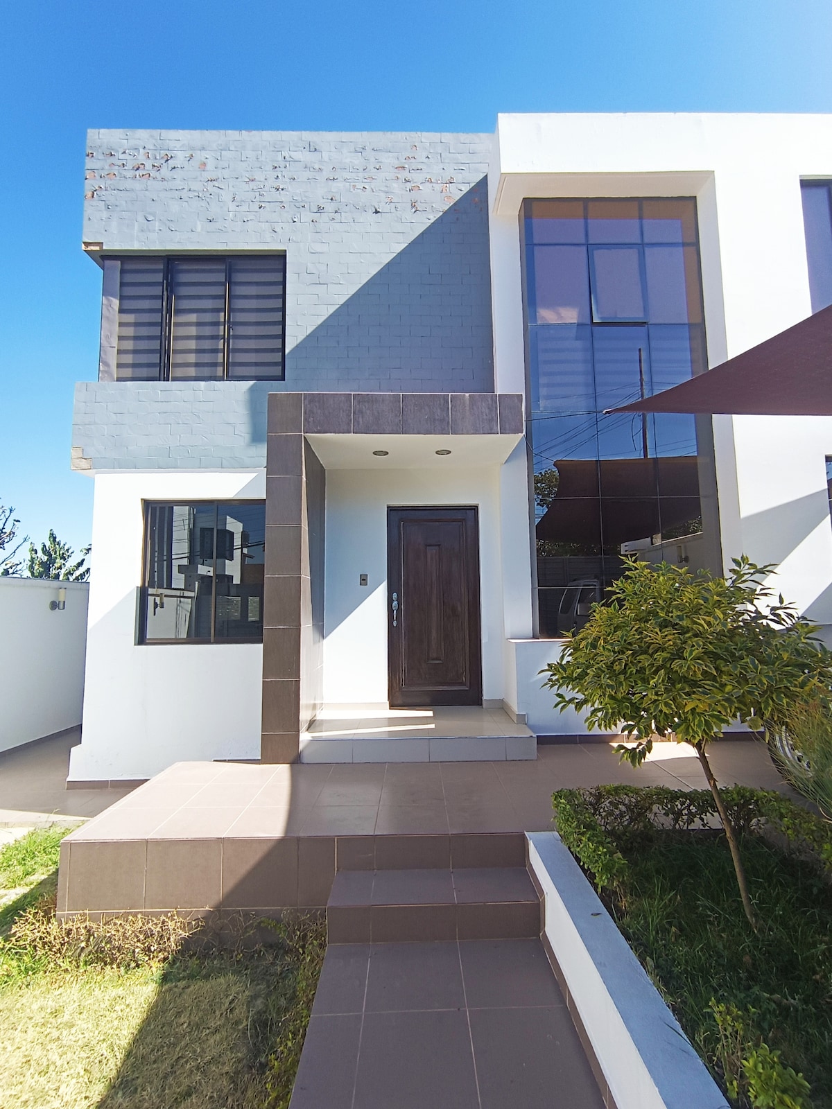 The exterior of a modern house is displayed, featuring large windows and a sleek entrance. A pathway leads to the front door, bordered by neatly trimmed grass and a small bush. Natural sunlight highlights the architectural details against a clear blue sky.