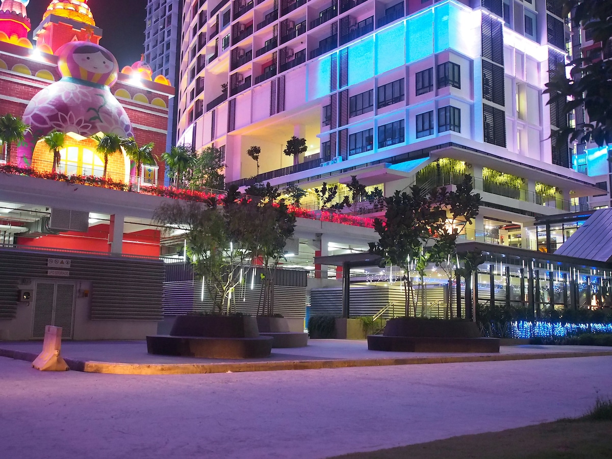 The vibrant exterior of the building is illuminated by colorful neon lights at night. Modern architecture is framed by lush greenery, with spacious seating areas visible on the ground level. The overall setting provides a lively and dynamic atmosphere, enhanced by artistic elements in the background.