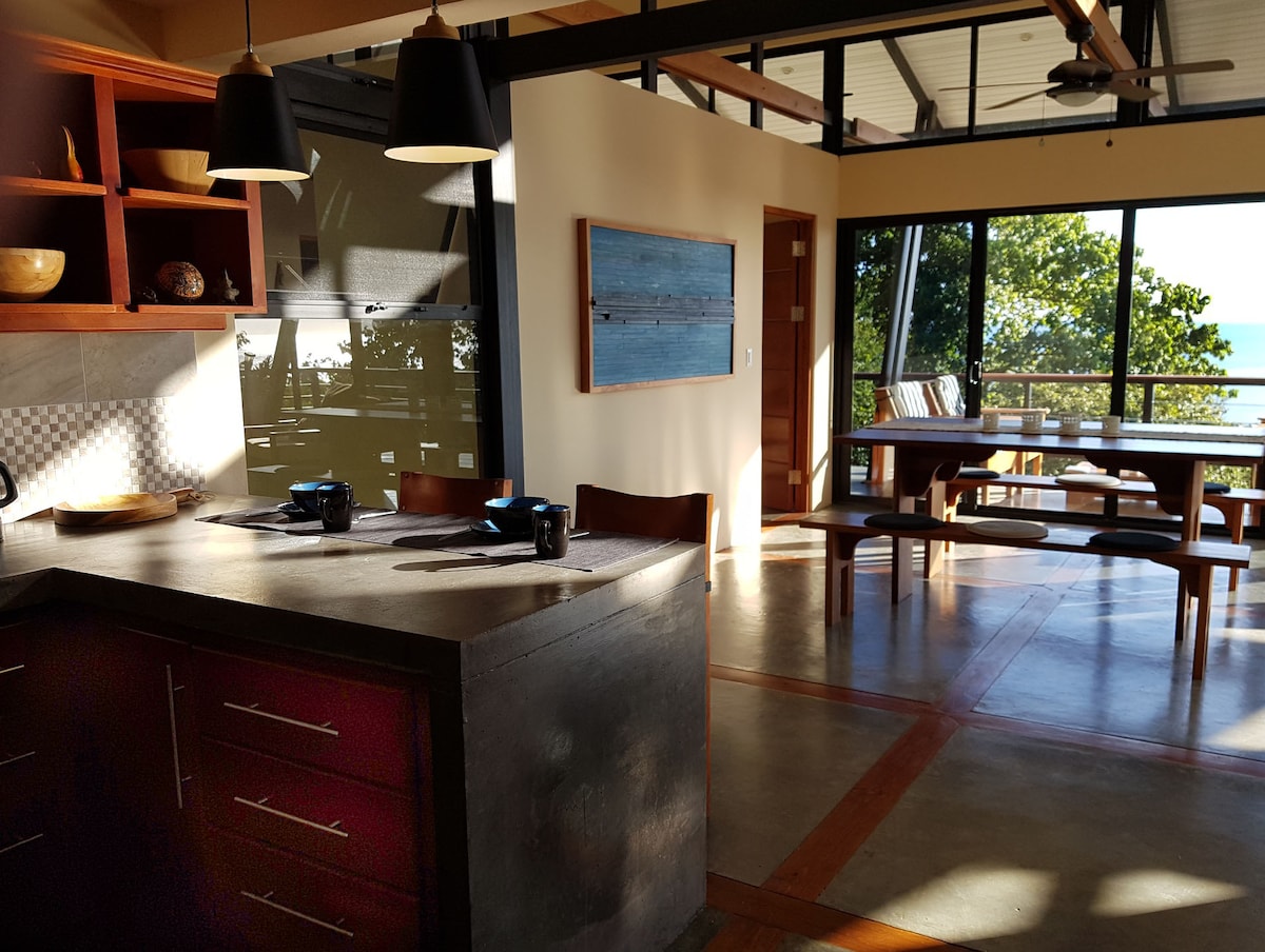 A modern kitchen island made of concrete features a sleek countertop. Wooden shelves display bowls and decorative items. A dining area with a large table and several chairs is visible, alongside expansive glass doors that offer views of the lush greenery outside.