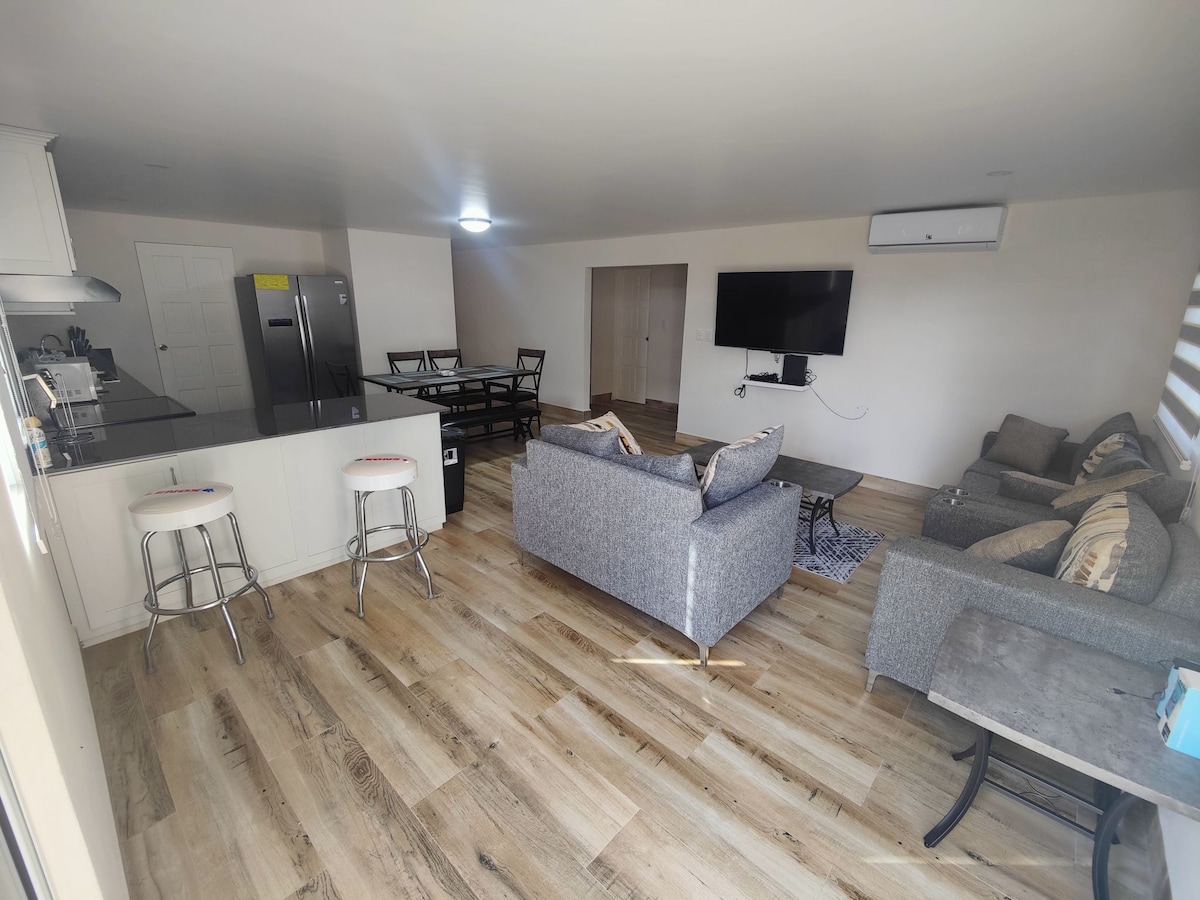 The living area presents an open layout featuring a comfortable seating arrangement with a sofa and armchair, complemented by a flat-screen television mounted on the wall. A modern kitchen island with bar stools is adjacent to a dining set, contributing to the inviting atmosphere.
