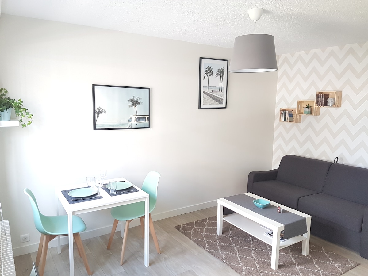 A bright living space is presented, featuring a small dining table with turquoise chairs set for two. A dark couch is positioned against the wall, complemented by a neutral rug. Artworks depicting palm trees adorn the walls, enhancing the airy atmosphere.