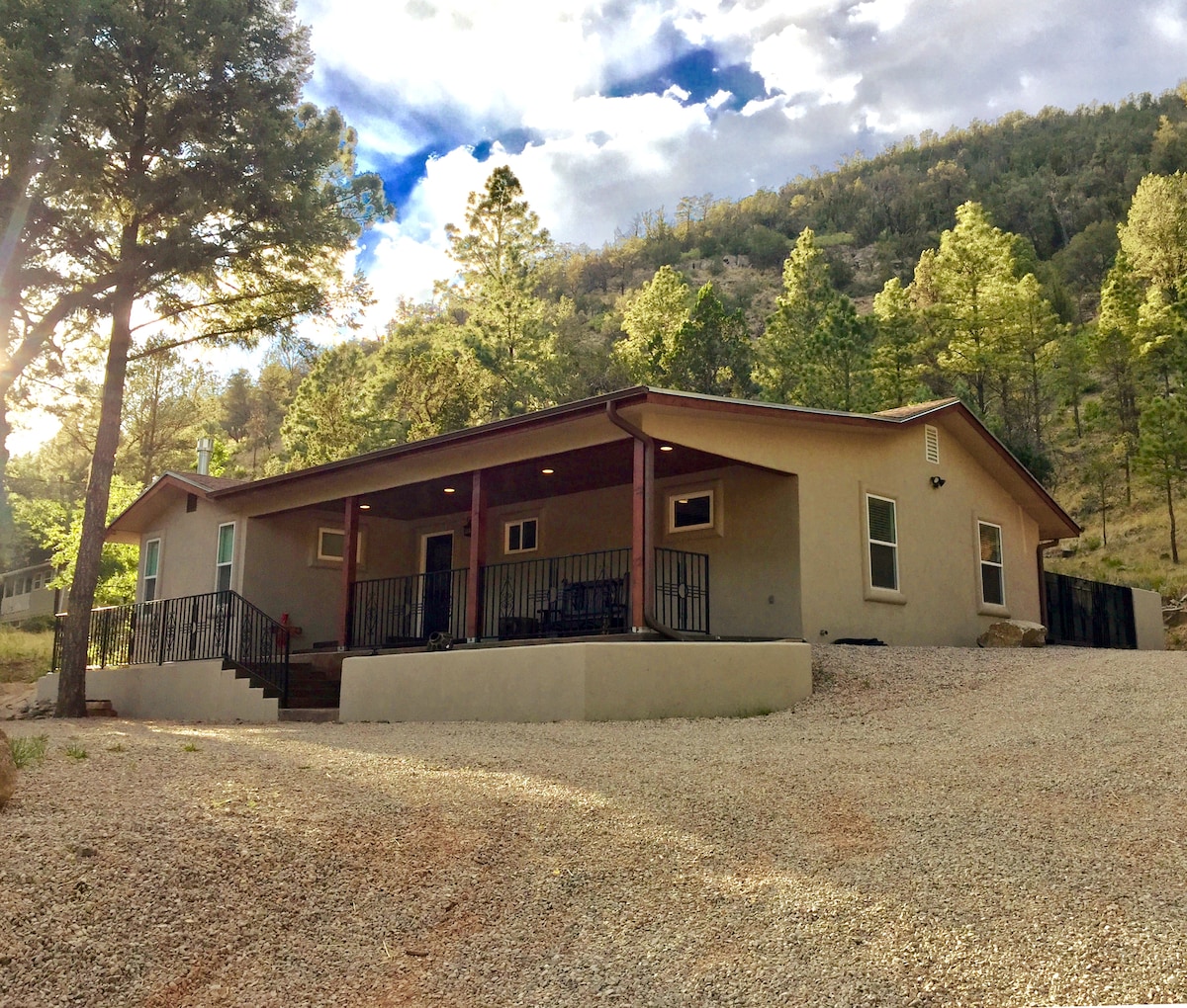 The exterior of a modern home is showcased, featuring a covered front porch with black railings. The house is surrounded by a gravel driveway and natural landscaping, including tall trees and mountain scenery in the background under a partly cloudy sky.