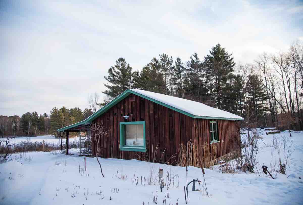 The cabin, featuring a rustic wooden exterior, is set against a backdrop of snow-covered fields and towering pine trees. A low profile roof complements the tranquil winter landscape, with a window visible on the side inviting natural light indoors.