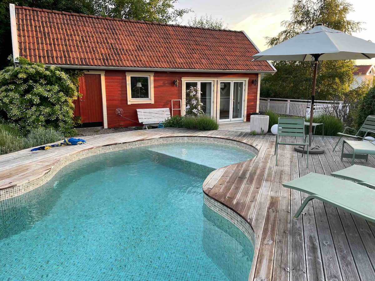 A serene pool area is framed by wooden decking, featuring a gently curving pool with clear blue water. A cozy red cabin is positioned in the background, complemented by green landscaping and a patio umbrella providing shade.