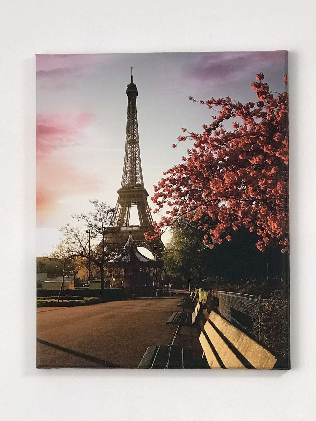 A canvas print displays the Eiffel Tower framed by blooming trees in vibrant pink. A pathway lined with benches leads to the tower, illuminated by soft evening light, creating a serene atmosphere in a Parisian setting.