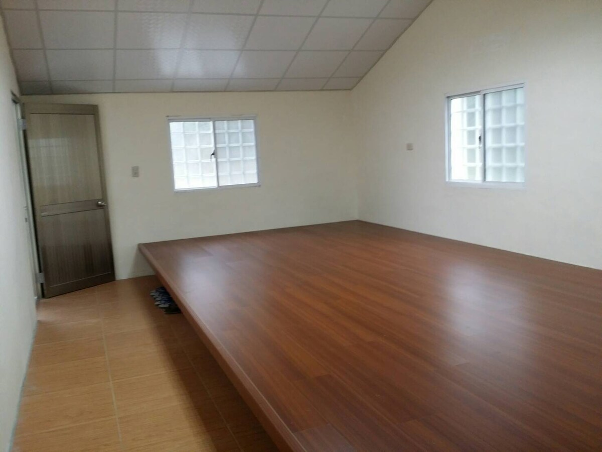 A spacious bedroom features a large wooden platform bed occupying the center. Two windows allow natural light to fill the room, surrounded by light-colored walls and a ceiling with a neutral finish. A door leads to an adjoining space.
