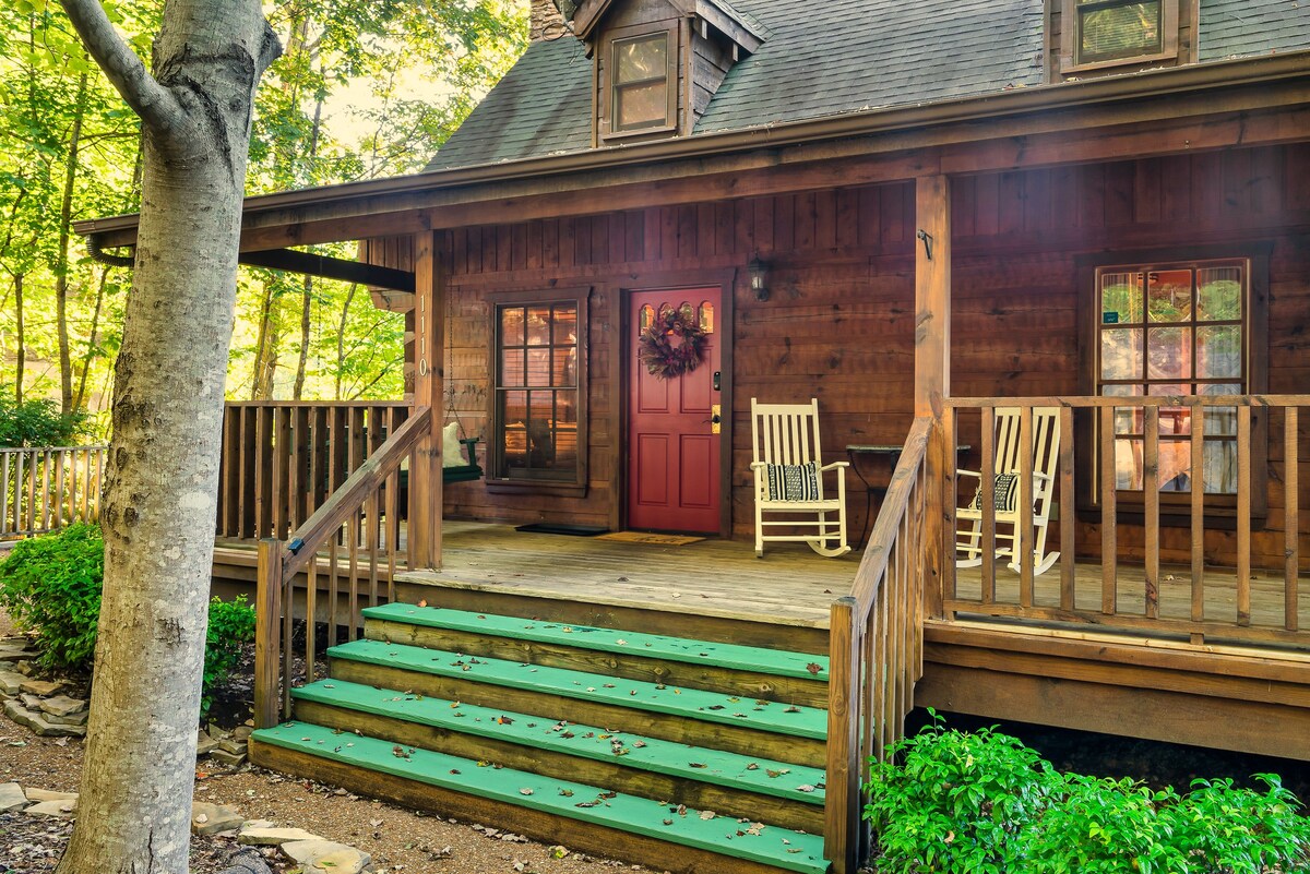 An inviting log cabin entrance features a red front door adorned with a wreath. Wooden steps lead up to a spacious porch, which includes rocking chairs and railing. Surrounding lush greenery complements the natural setting, enhancing the serene atmosphere of the cabin.