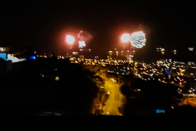 The night scene captures a vibrant display of fireworks illuminating the sky above a cityscape. Twinkling lights from streets and buildings create a warm glow, while the expansive view showcases the coastal surroundings and distant hills.