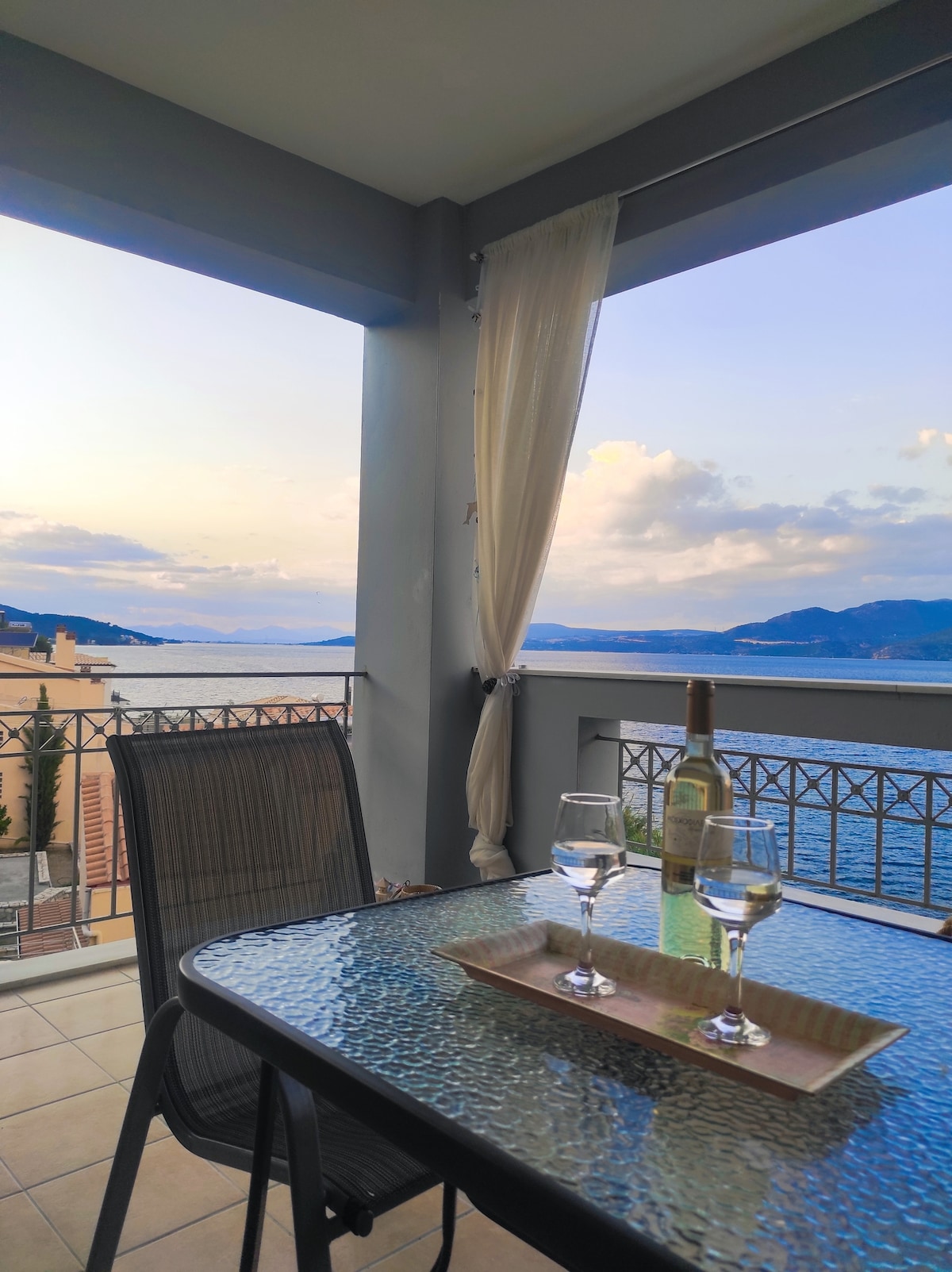 A terrace is seen featuring a glass-top table with two wine glasses and a bottle of wine. A light curtain billows gently in the breeze, framing a scenic view of the Ionian Sea and distant mountains under a pastel sky.